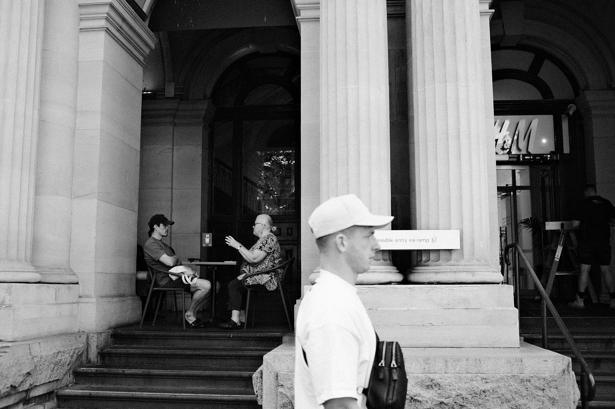 Couple in conversation under columns, stranger passing in foreground
